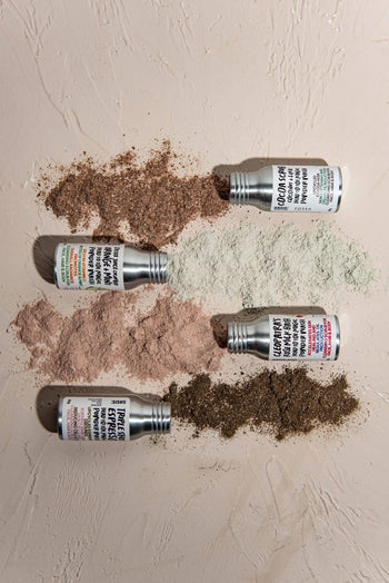 Four small containers of Oasis Beauty Kitchen Head to Toe Magic Powder Wash, 25g, are spilled on a light textured surface, their vegan, plant-based powders in brown, green, pink, and dark brown forming colored piles with labels visible.