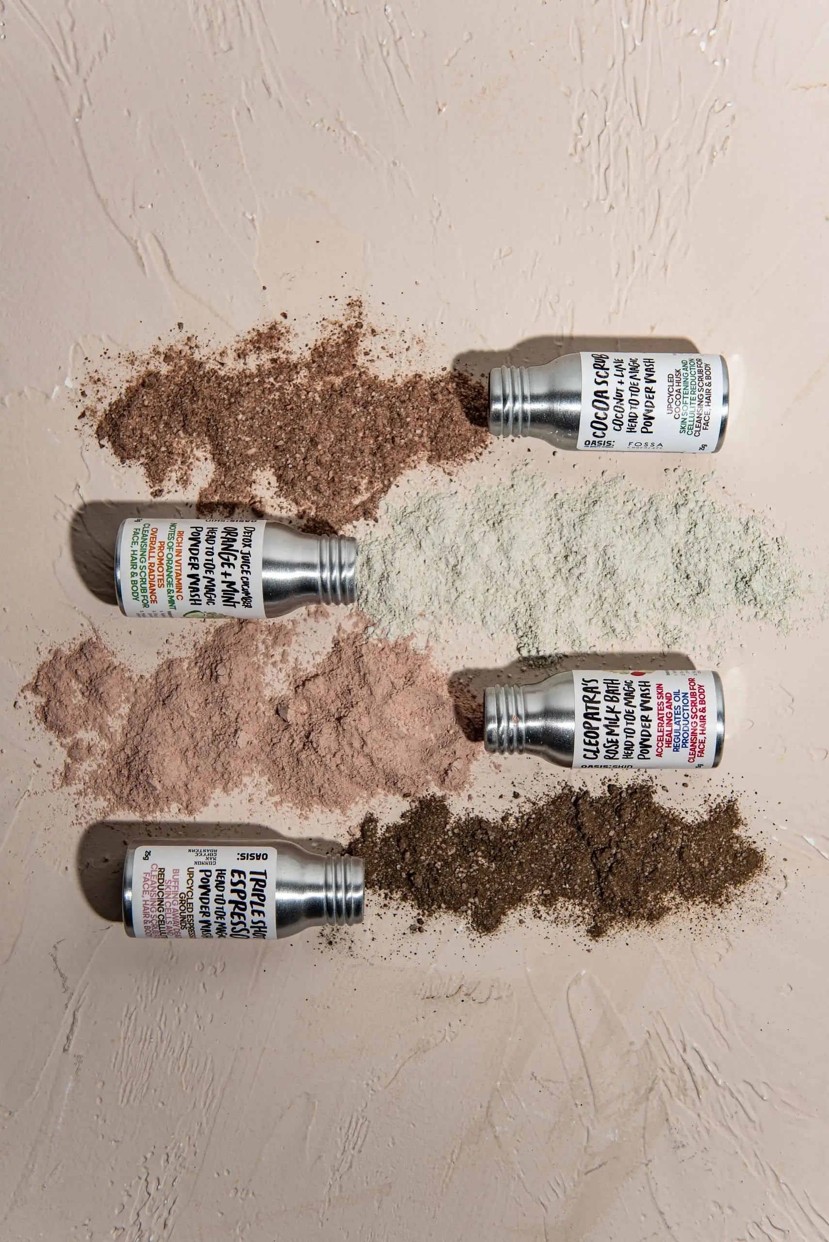 Four bottles of Oasis Beauty Kitchen Head to Toe Magic Powder Wash, 25g, from Oasis Beauty Kitchen, lie tipped on a textured surface, spilling brown, green, pink, and dark brown plant-based powders in horizontal rows.