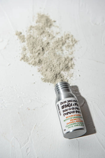 A small bottle labeled Oasis Beauty Kitchen Head to Toe Magic Powder Wash, 25g, rests on a white surface with some light gray powder spilled—crafted by Oasis Beauty Kitchen for a gentle, effective vegan wash using plant-based ingredients.
