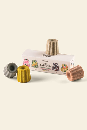 Three colorful, canelé-shaped Oasis Beauty Kitchen Miniature Solid Shampoos sit beside and atop a rectangular box labeled The Classics from Oasis Beauty Kitchen. The box shows illustrations that match the gray, yellow, brown, and tan shampoos.