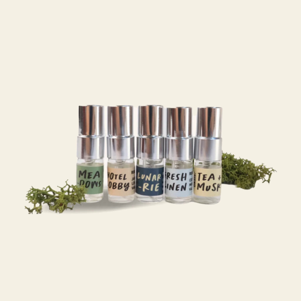 The Oasis Beauty Kitchen Miniscapes Scent Discovery Kit features six small glass bottles with silver caps—MEAD DOWS, HOTEL LOBBY, LUNAR RIES, FRESH LINEN, and TEA + MUSK—arranged in a row on a light background with sprigs of moss.