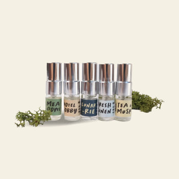 The Oasis Beauty Kitchen Miniscapes Scent Discovery Kit features six small glass bottles with silver caps—MEAD DOWS, HOTEL LOBBY, LUNAR RIES, FRESH LINEN, and TEA + MUSK—arranged in a row on a light background with sprigs of moss.