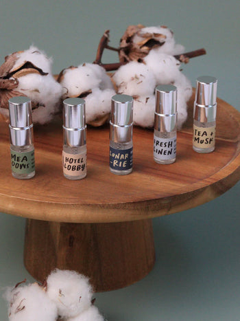 Five mini bottles from Oasis Beauty Kitchen’s Miniscapes Scent Discovery Kit—Meadows, Hotel Lobby, Lunar Rie, Fresh Linen, and Tea + Musk—sit on a wooden pedestal surrounded by cotton branches. Ideal for scent lovers seeking an aromatic escape.