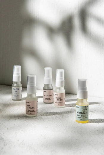 Five small Oasis Beauty Kitchen Room and Linen Mist (10ml) bottles are arranged on a white textured surface with soft natural light and leaf shadows. The yellow-labeled front bottle highlights the Fresh Linen scent.