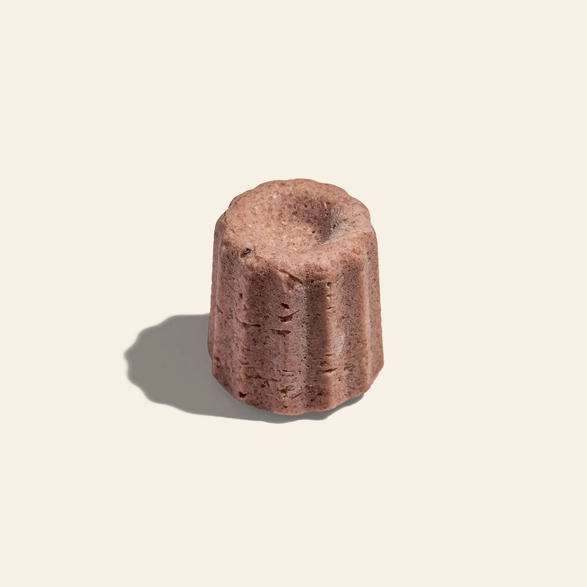 A single Oasis Beauty Kitchen Rose Solid Shampoo, Midi Canele, from Oasis Beauty Kitchen, with a fluted cylindrical shape and caramelized exterior, is delicately garnished with Damascus Rose Petals on a plain light background.