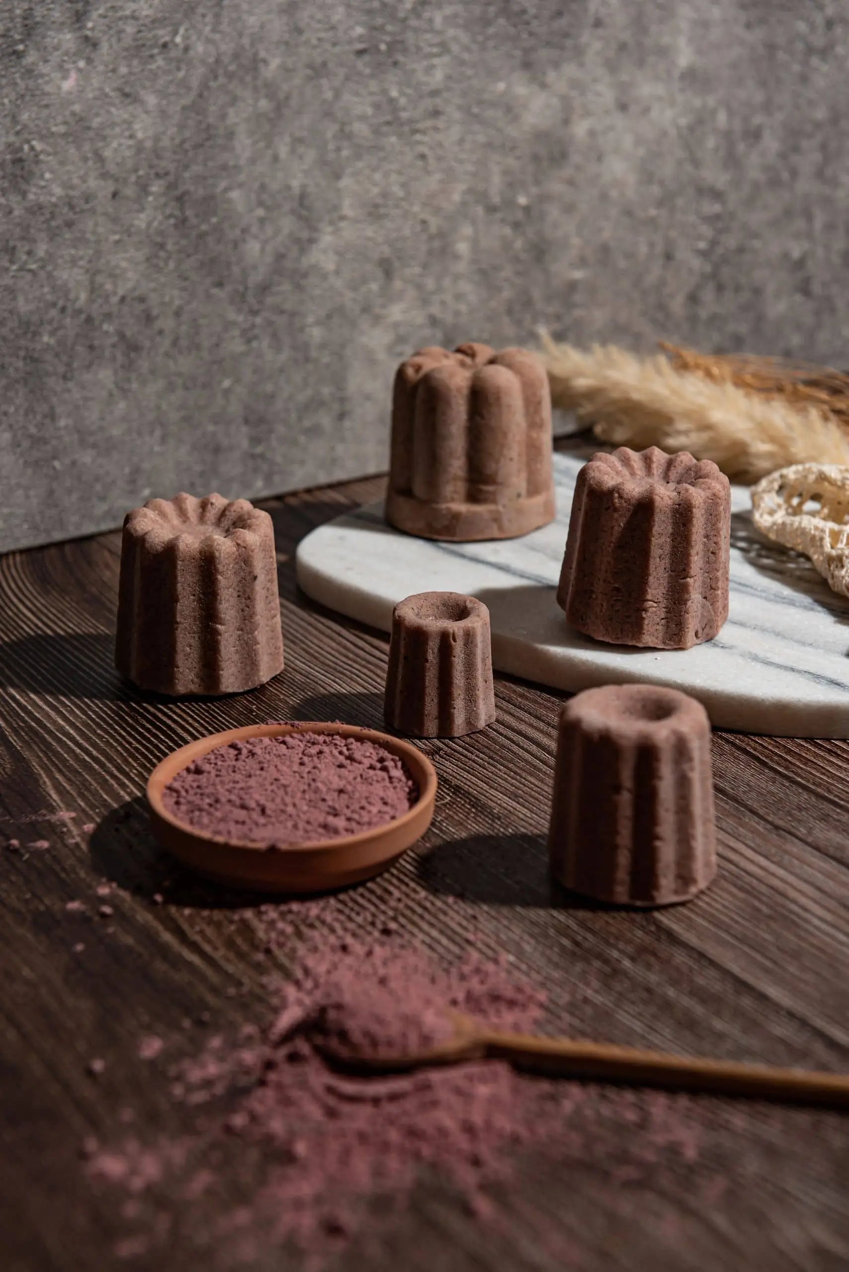 Oasis Beauty Kitchen Rose Solid Shampoo, Midi Canele by Oasis Beauty Kitchen is displayed on a dark wooden surface and marble board, surrounded by a small dish of pink powder, a wooden spoon, dried plants, and delicate Damascus Rose Petals.