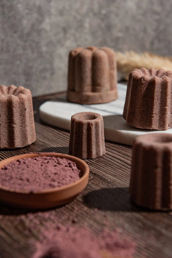 Oasis Beauty Kitchen Rose Solid Shampoo Midi Canele by Oasis Beauty Kitchen is displayed on wood and marble, with a shallow cocoa powder dish and organic rosemary in front. The softly blurred background highlights the elegant arrangement.