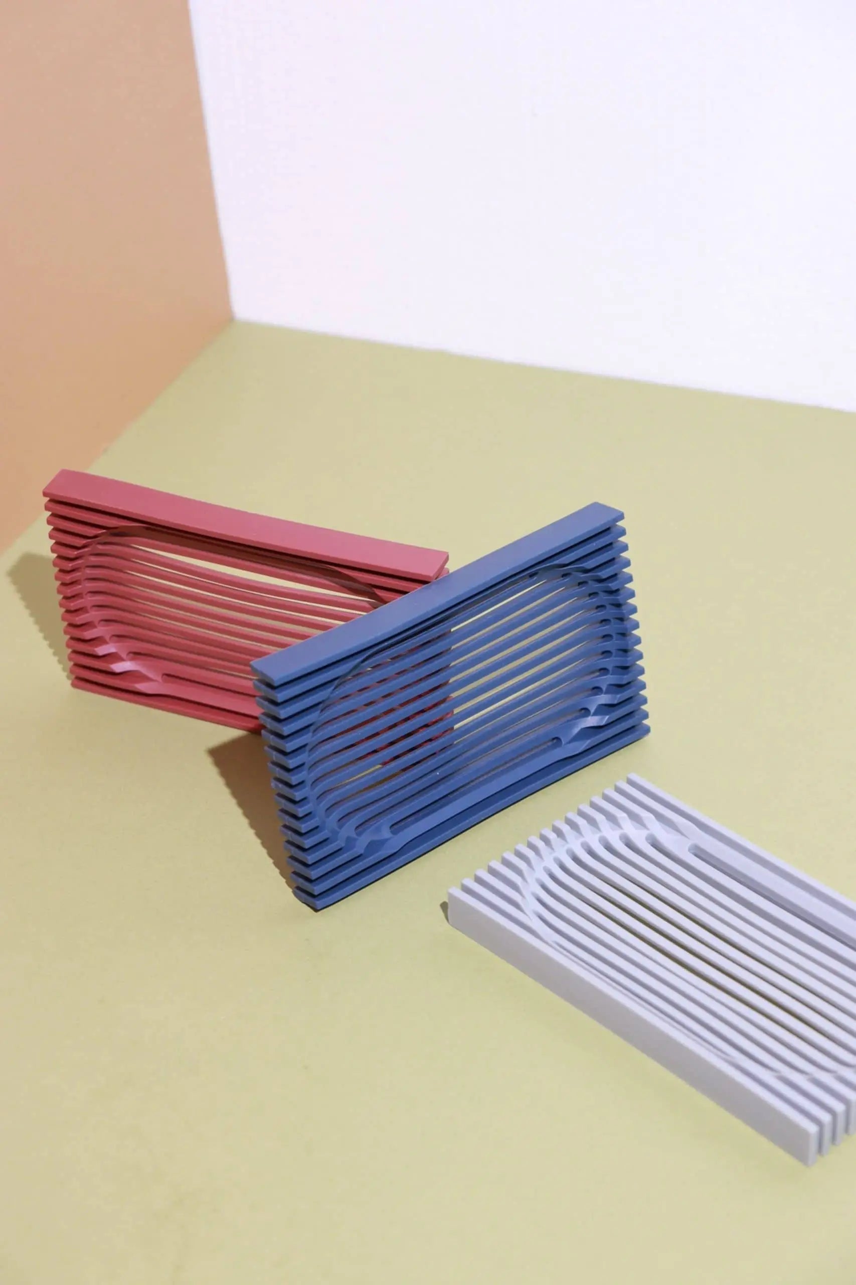 Three Oasis Beauty Kitchen The Lattice Soap Dishes by Oasis Beauty Kitchen in red, blue, and white are arranged on a light green surface with a peach and white background, highlighting their minimalist geometric design and sturdy construction.