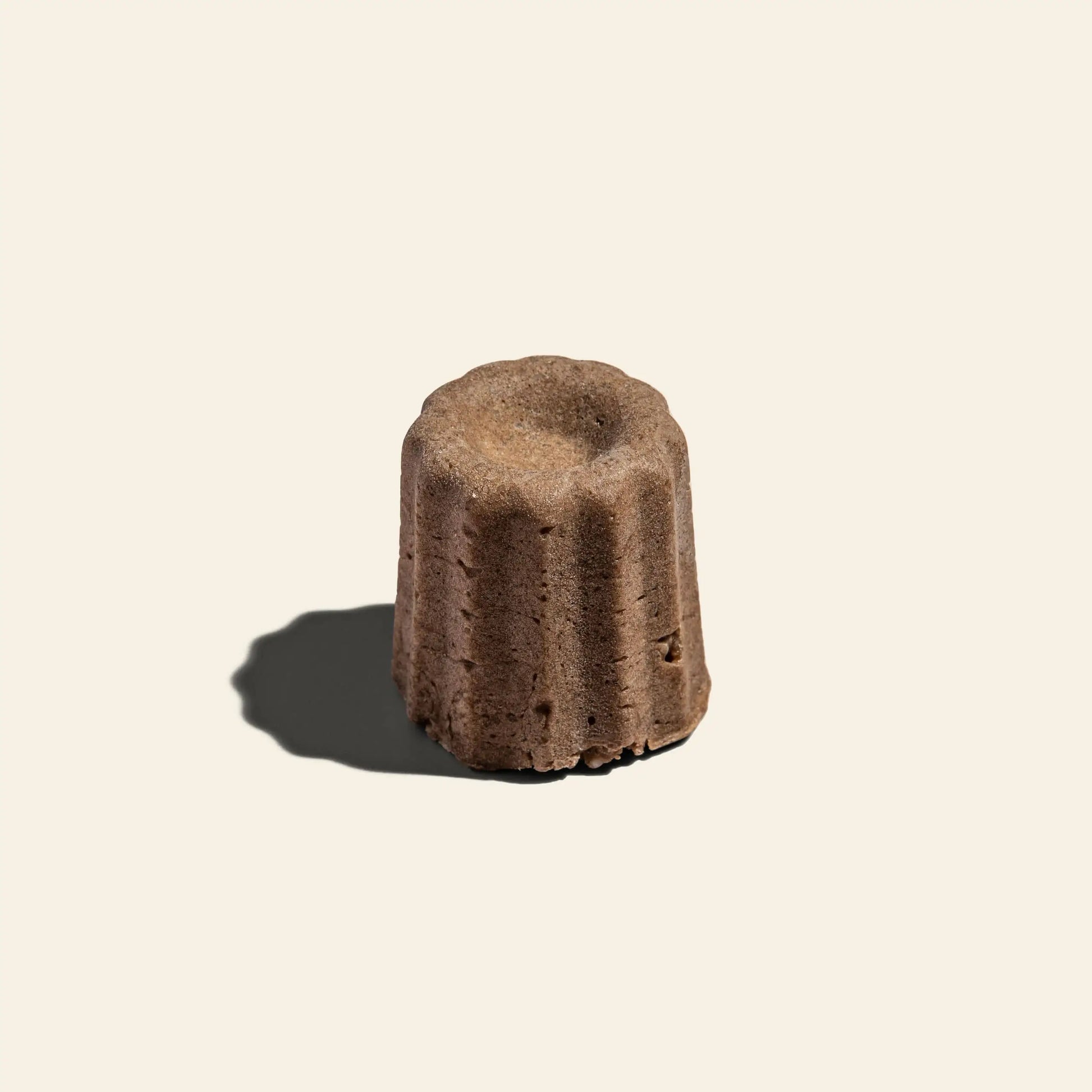 A single Oasis Beauty Kitchen Uji Hojicha Solid Shampoo, Midi Canele, with a fluted brown form, sits on a light beige background dusted with Hojicha powder and casting a leftward shadow.