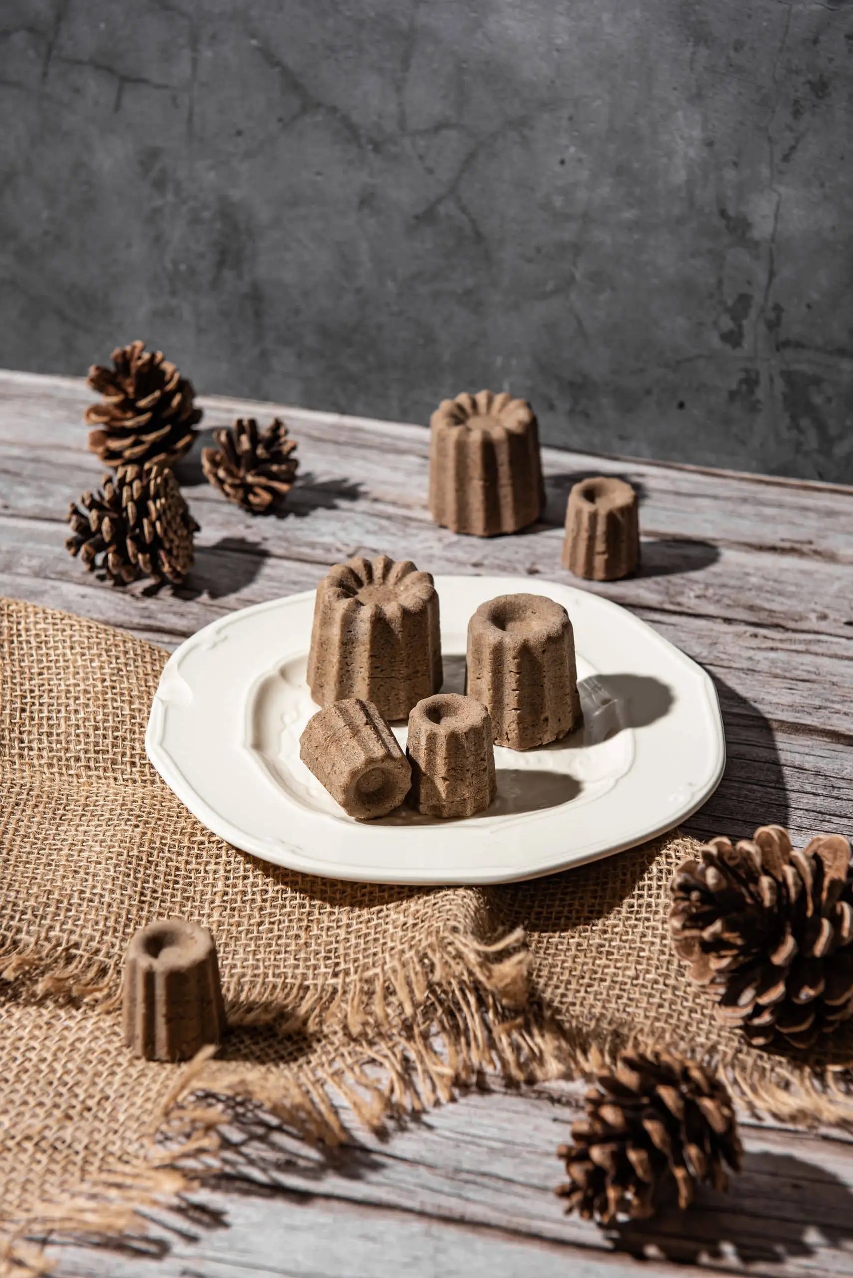 A white plate displays several brown, canelé-shaped Oasis Beauty Kitchen Uji Hojicha Solid Shampoo bars, each dusted with Hojicha powder, set on a rustic wooden table adorned with burlap and pinecones.