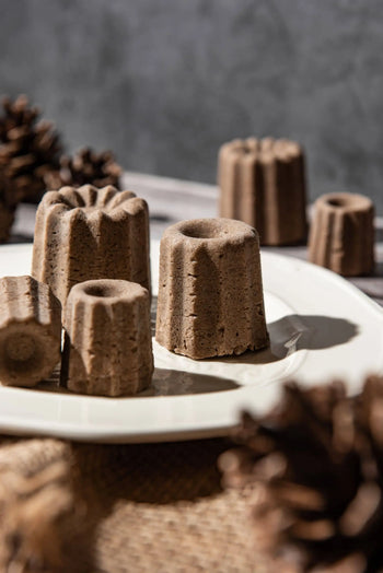 A white plate displays several brown, canelé-shaped Oasis Beauty Kitchen Uji Hojicha Solid Shampoo bars, crafted with Hojicha powder, with blurred pinecones in the background.