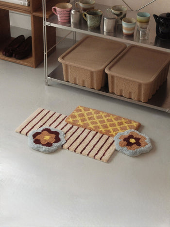 Three colorful, textured rugs—including the Ordinary Daily Design Deconstructed: Flower Rug in a soft pink asymmetrical flower shape, one striped rug, and another with a diamond pattern—are placed on a smooth floor beside a metal shelf that holds cups, trays, and a tea kettle.