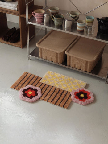 Two Ordinary Daily Design Deconstructed: Flower Rugs by Ordinary Daily Design and an asymmetrical pink striped rug are arranged on a light floor next to a metal shelf displaying cups, mugs, and beige storage bins.