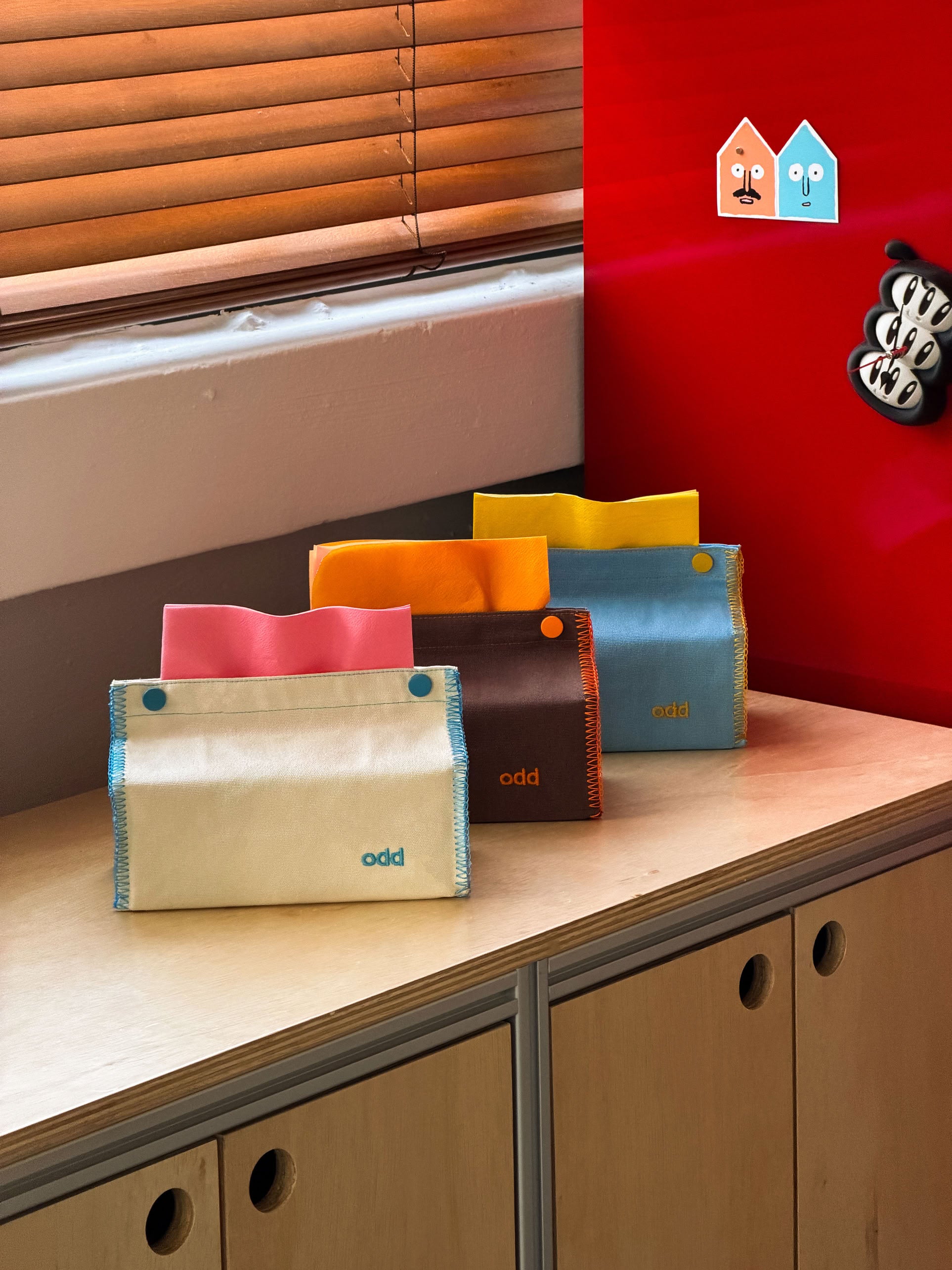 Four colorful Ordinary Daily Design Tissue Cases with the word “odd” are displayed on a wooden cabinet, each resembling a stylish fabric cover with blue and yellow stitching from Ordinary Daily Design. In the background, a red wall, wooden blinds, and two small house-shaped decorations complete the cozy scene—making each tissue case both playful and practical for your space.
