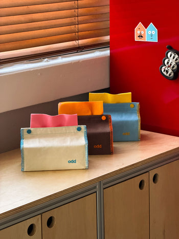 Four colorful Ordinary Daily Design Tissue Cases with the word “odd” are displayed on a wooden cabinet, each resembling a stylish fabric cover with blue and yellow stitching from Ordinary Daily Design. In the background, a red wall, wooden blinds, and two small house-shaped decorations complete the cozy scene—making each tissue case both playful and practical for your space.