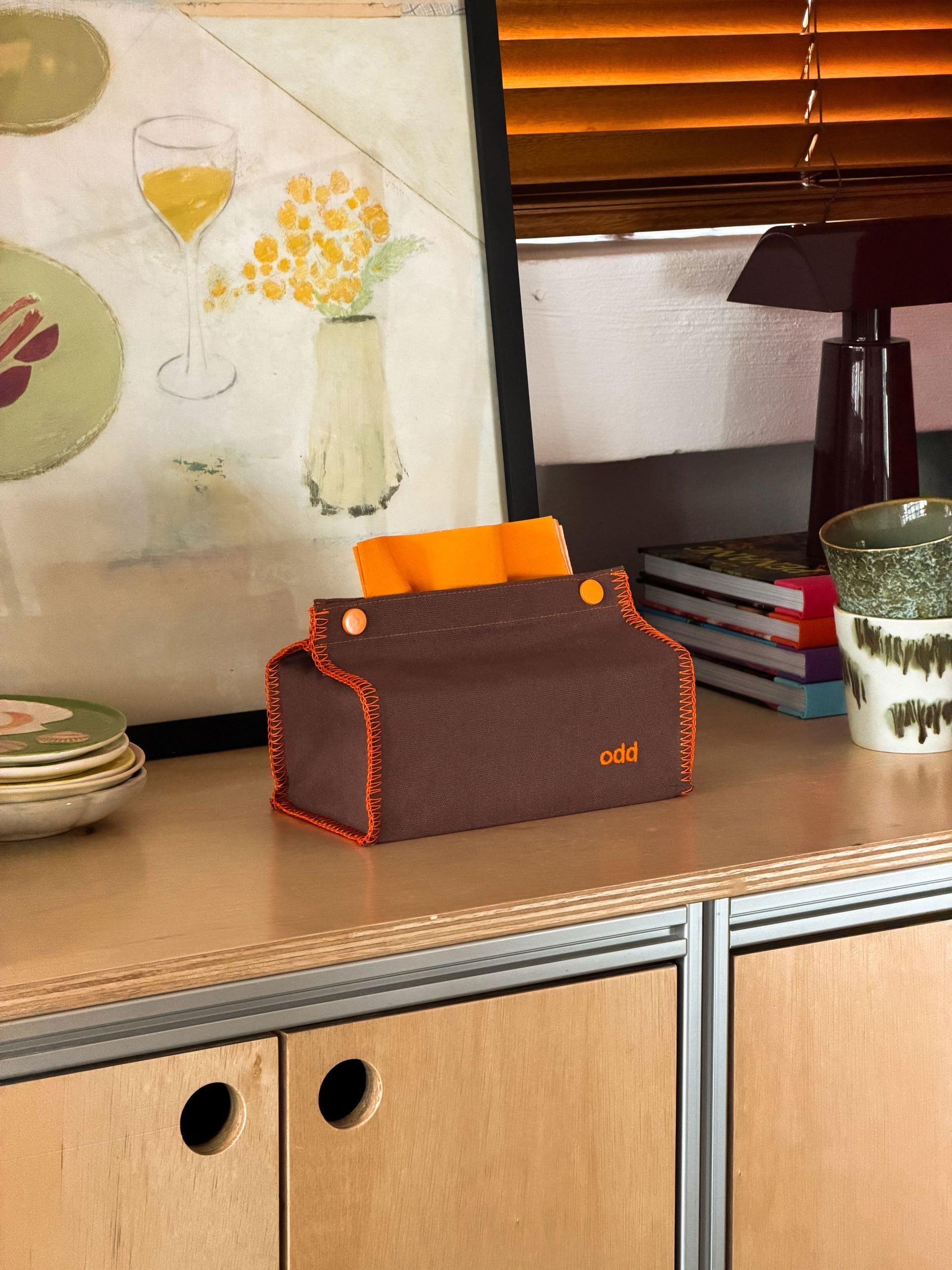 An Ordinary Daily Design Tissue Case in brown with orange stitching, labeled “odd,” rests on a wooden cabinet beside stacked books, a cup, and dishes, with orange blinds and artwork in the background.