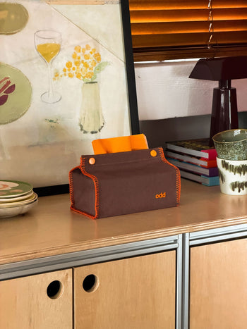 An Ordinary Daily Design Tissue Case in brown with orange stitching, labeled “odd,” rests on a wooden cabinet beside stacked books, a cup, and dishes, with orange blinds and artwork in the background.
