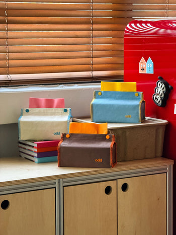 Four colorful fabric pouches, including the Ordinary Daily Design Tissue Case in Cream/Blue Stitch from Ordinary Daily Design with decorative button accents, sit on a wooden cabinet near a red fridge, stacked on books and a box, with wooden blinds in the background.