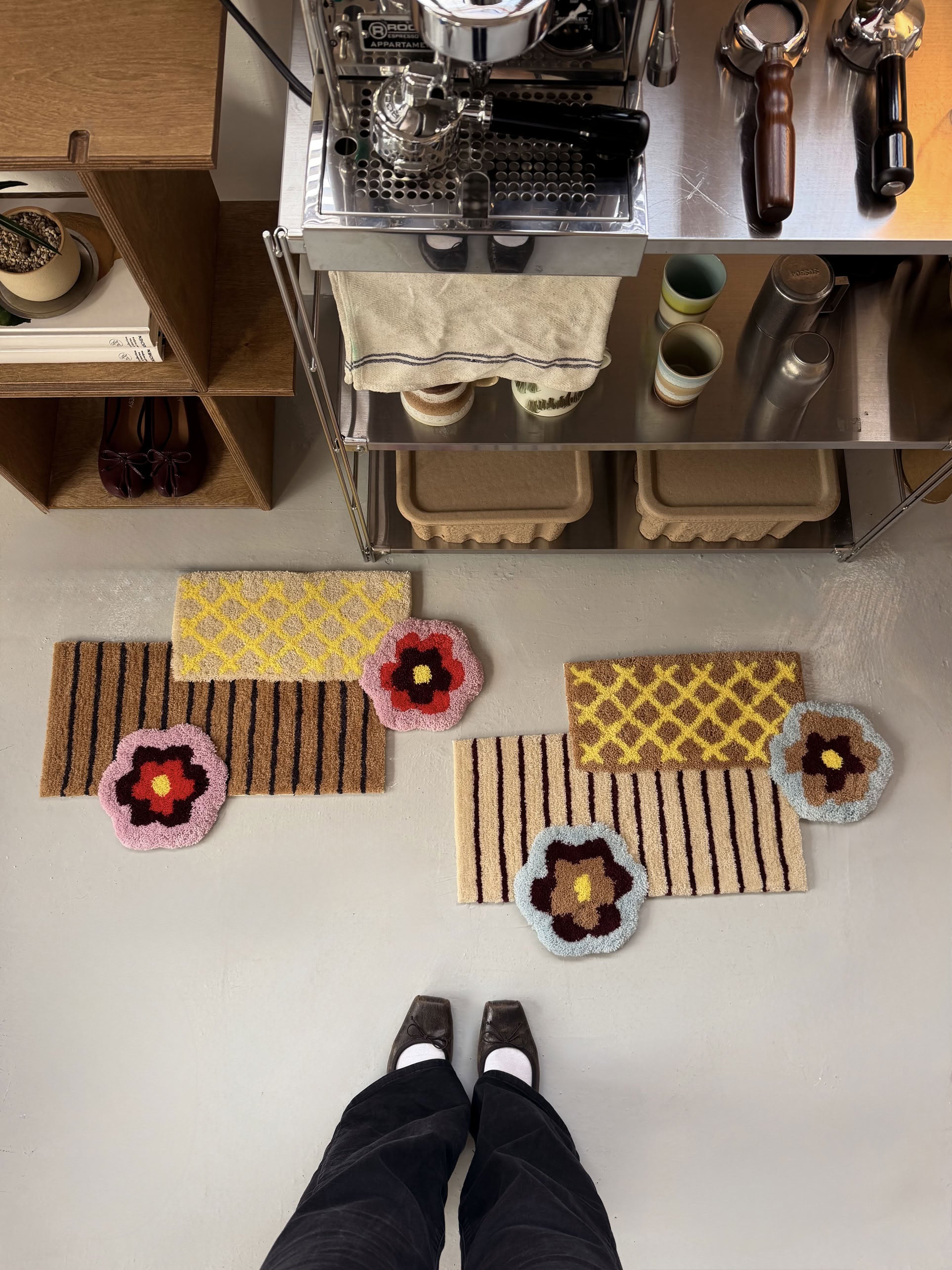 A person stands in front of a coffee station with mugs and equipment. On the floor are colorful, patterned rugs—including a pink rug and the Ordinary Daily Design Deconstructed: Flower Rug by Ordinary Daily Design, which features bold floral and geometric patterns. Only the person’s legs and shoes are visible.