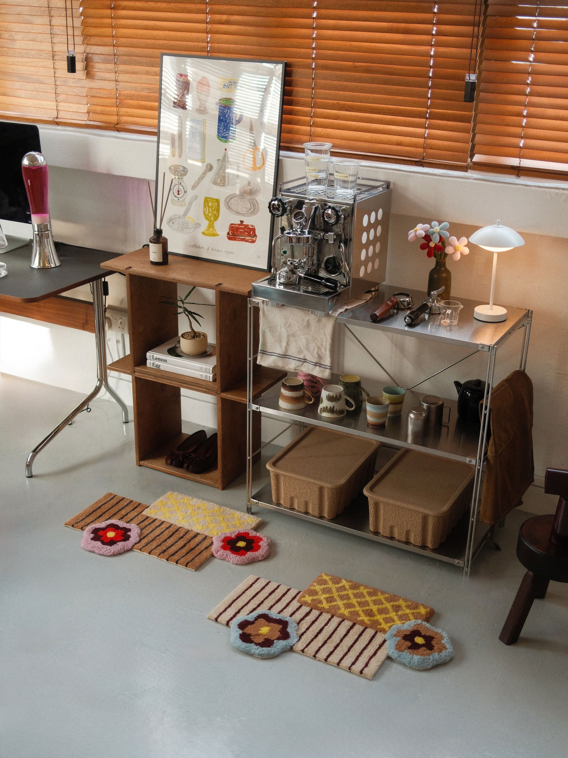 A cozy corner features a coffee machine, cups, lamp, decorative items, and an art print displayed on shelves; two woven baskets sit underneath; the Ordinary Daily Design Deconstructed: Flower Rug from Ordinary Daily Design adds a vibrant touch to the floor; blinds cover the window behind.