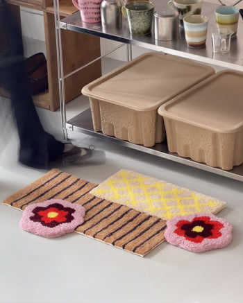 A person walks past three colorful rugs—one with stripes, one featuring a yellow lattice pattern, and the Ordinary Daily Design Deconstructed: Flower Rug by Ordinary Daily Design in striking red and pink—placed in front of shelves holding beige bins.