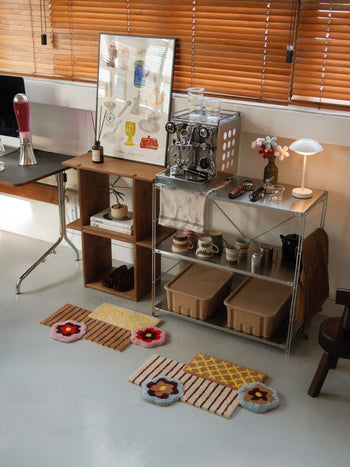 A cozy home coffee station featuring an espresso machine, cups, shelves with baskets and mugs, a framed art print, and the Ordinary Daily Design Deconstructed: Flower Rug by Ordinary Daily Design on the floor. Blinds cover the window, and a lava lamp sits on the desk.