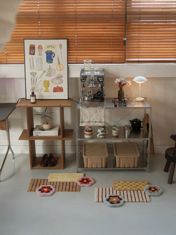 A cozy corner featuring the Ordinary Daily Design Deconstructed: Flower Rug by Ordinary Daily Design in soft hues on a white floor, complemented by a coffee machine on a clear cart, cups, a small lamp, an art print, wooden shelf, shoes, books, and wooden blinds in the background.