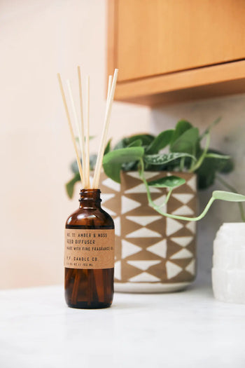 The P.F. Candle Co. 3.5oz Reed Diffuser in Amber & Moss, with wooden reeds and brown glass bottle, sits on a white countertop next to a potted plant and textured container, beneath warm wooden cabinets.