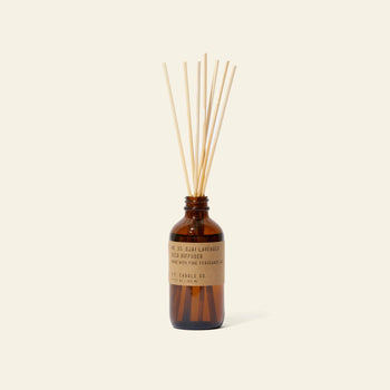 The P.F. Candle Co. 3.5oz Reed Diffuser in Ojai Lavender features several natural reeds in a brown glass bottle with a tan label, releasing soothing fragrance oils into the air against a plain light background.