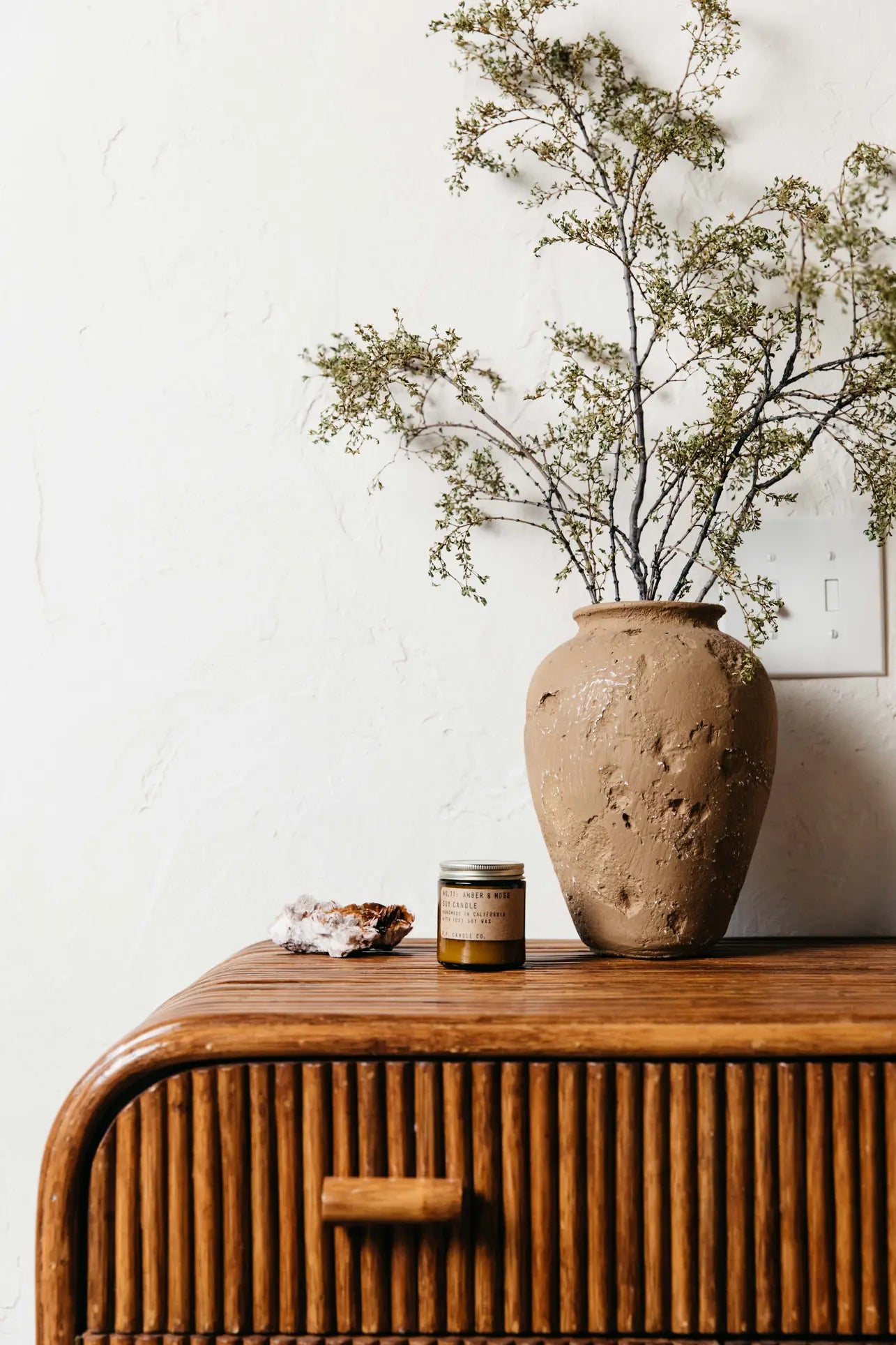 A textured clay vase with leafy branches and lavender sits on a wooden rattan dresser next to a P.F. Candle Co. No. 11 Amber & Moss 3.5 oz Mini Candle and a crystal cluster, all set against a white wall.