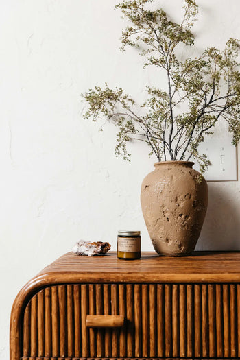 A textured clay vase with leafy branches and lavender sits on a wooden rattan dresser next to a P.F. Candle Co. No. 11 Amber & Moss 3.5 oz Mini Candle and a crystal cluster, all set against a white wall.