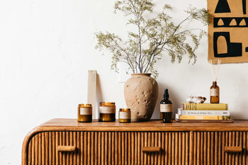 A wooden sideboard with amber glass jars, the P.F. Candle Co. No. 11 Amber & Moss 3.5 oz Mini Candle, a beige ceramic vase with greenery, a glass spray bottle, books, and a reed diffuser creates a minimal and cozy decor scene.