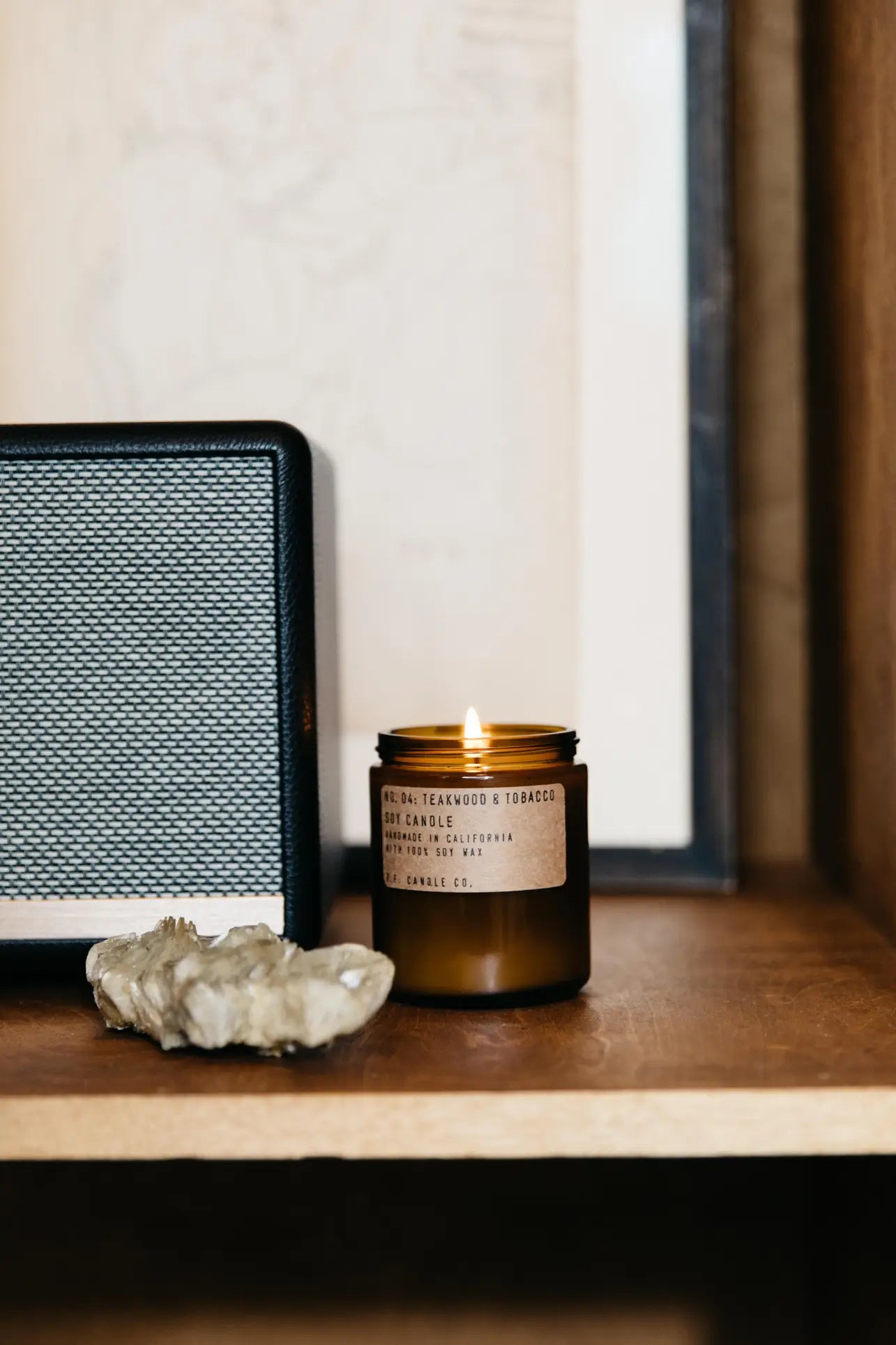 A P.F. Candle Co. No. 4 Teakwood & Tobacco 7.2 oz Soy Candle sits on a wooden shelf next to a black-and-silver speaker and crystals, with framed art in the background.