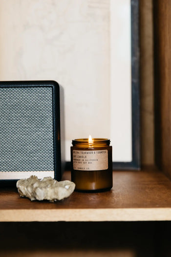 A P.F. Candle Co. No. 4 Teakwood & Tobacco 7.2 oz Soy Candle sits on a wooden shelf next to a black-and-silver speaker and crystals, with framed art in the background.
