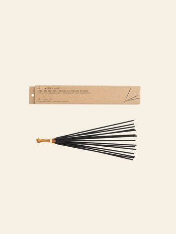 A bundle of black incense sticks tied with a tan string rests below a rectangular P.F. Candle Co. Classic Incense package labeled No. 11 Amber & Moss, evoking subtle notes of teakwood on a simple light background.
