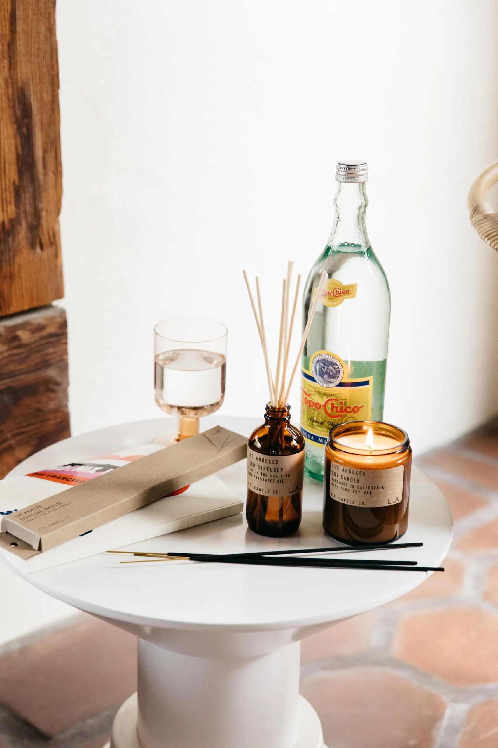 A small white round table with a glass of water, a bottle of Topo Chico, the P.F. Candle Co. Los Angeles Limited Edition 7.2 oz Soy Candle, and incense sticks brings inviting California vibes to this rustic indoor space.