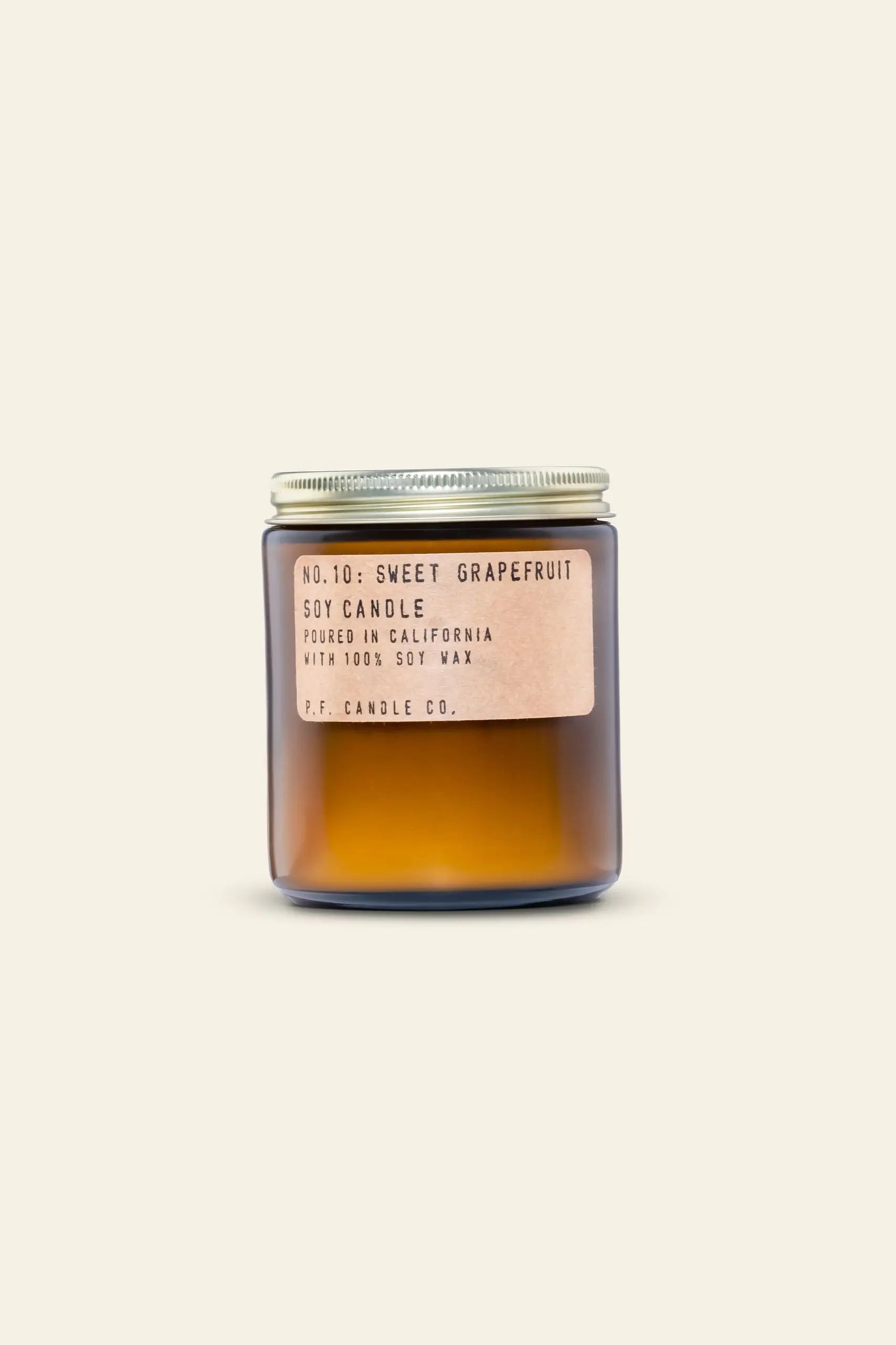 The P.F. Candle Co. No. 10 Sweet Grapefruit 7.2 oz Soy Candle, in brown glass with a silver lid, fills the air with a refreshing grapefruit scent against a light beige background.