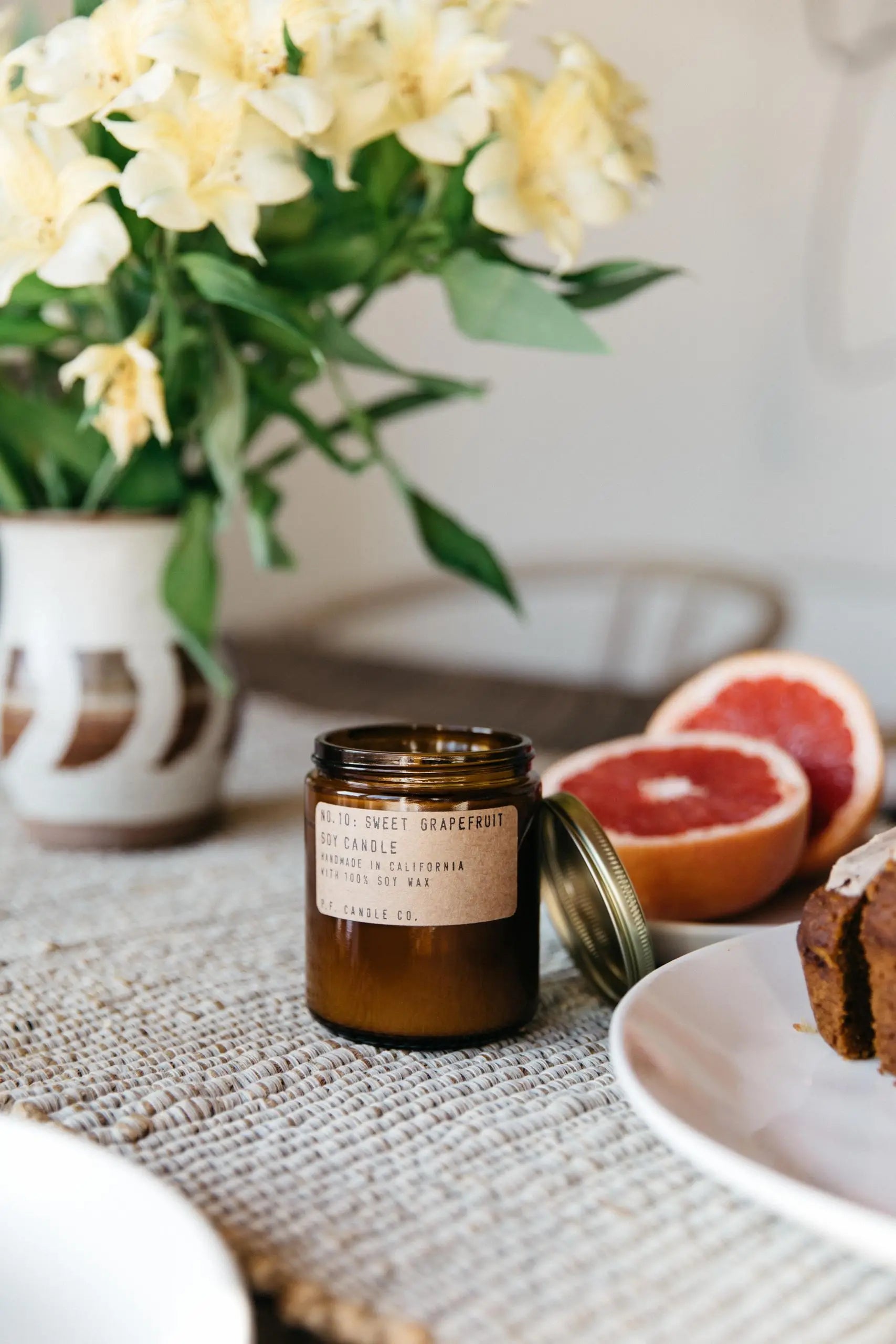 A P.F. Candle Co. No. 10 Sweet Grapefruit 7.2 oz Soy Candle sits on a woven placemat with its lid off, surrounded by grapefruit halves, white flowers in a vase, and a plate with a baked good—bringing refreshing vibes to any space.