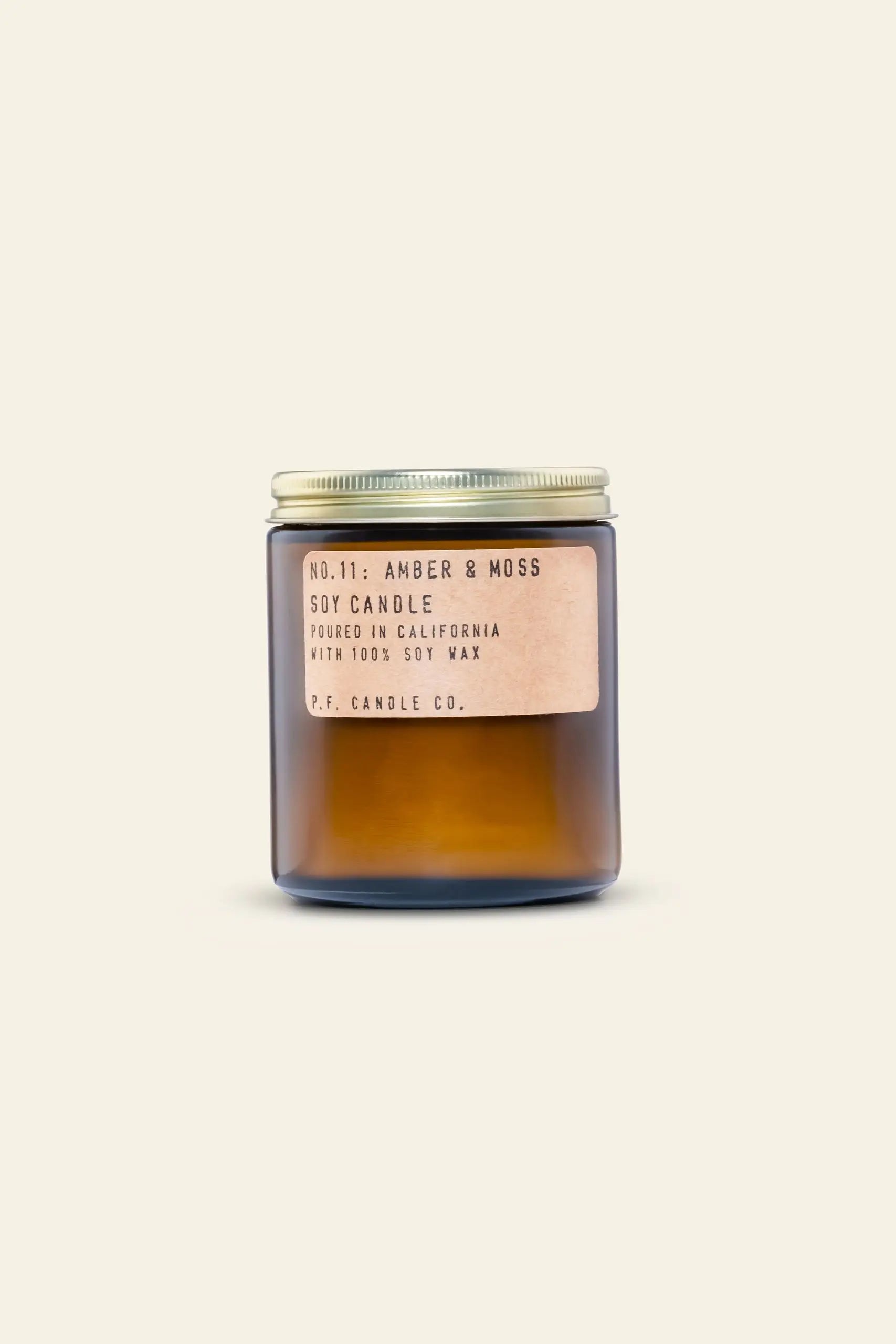 A 7.2 oz amber glass jar with a metal lid and beige label says P.F. Candle Co. No. 11 Amber & Moss Soy Candle, poured in California with all-natural soy wax for a calming aroma, set against a light background.