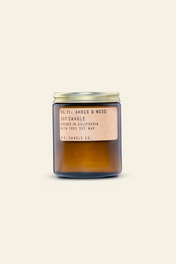 A 7.2 oz amber glass jar with a metal lid and beige label says P.F. Candle Co. No. 11 Amber & Moss Soy Candle, poured in California with all-natural soy wax for a calming aroma, set against a light background.