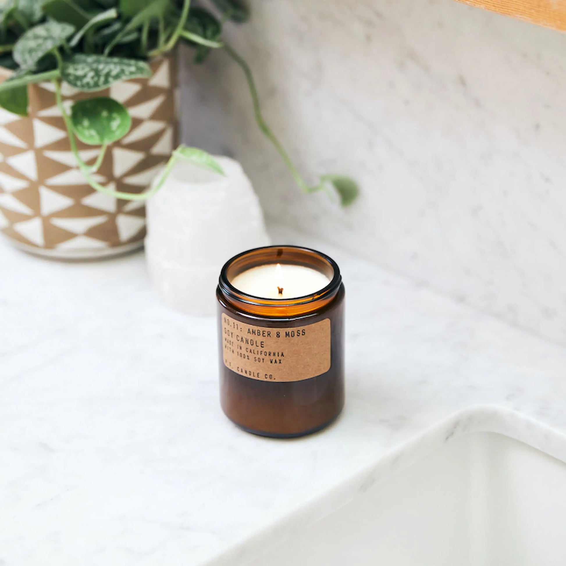 The P.F. Candle Co. No. 11 Amber & Moss 7.2 oz Soy Candle, in a brown glass jar, sits on a white marble countertop beside a patterned plant pot and white crystal, filling the space with a calming aroma.
