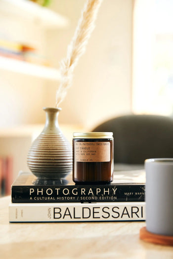 A P.F. Candle Co. No. 19 Patchouli Sweetgrass 7.2 oz Soy Candle rests on two books beside a striped ceramic vase with pampas grass; a gray mug is partially visible, creating an inviting scene for relaxation and reflection.