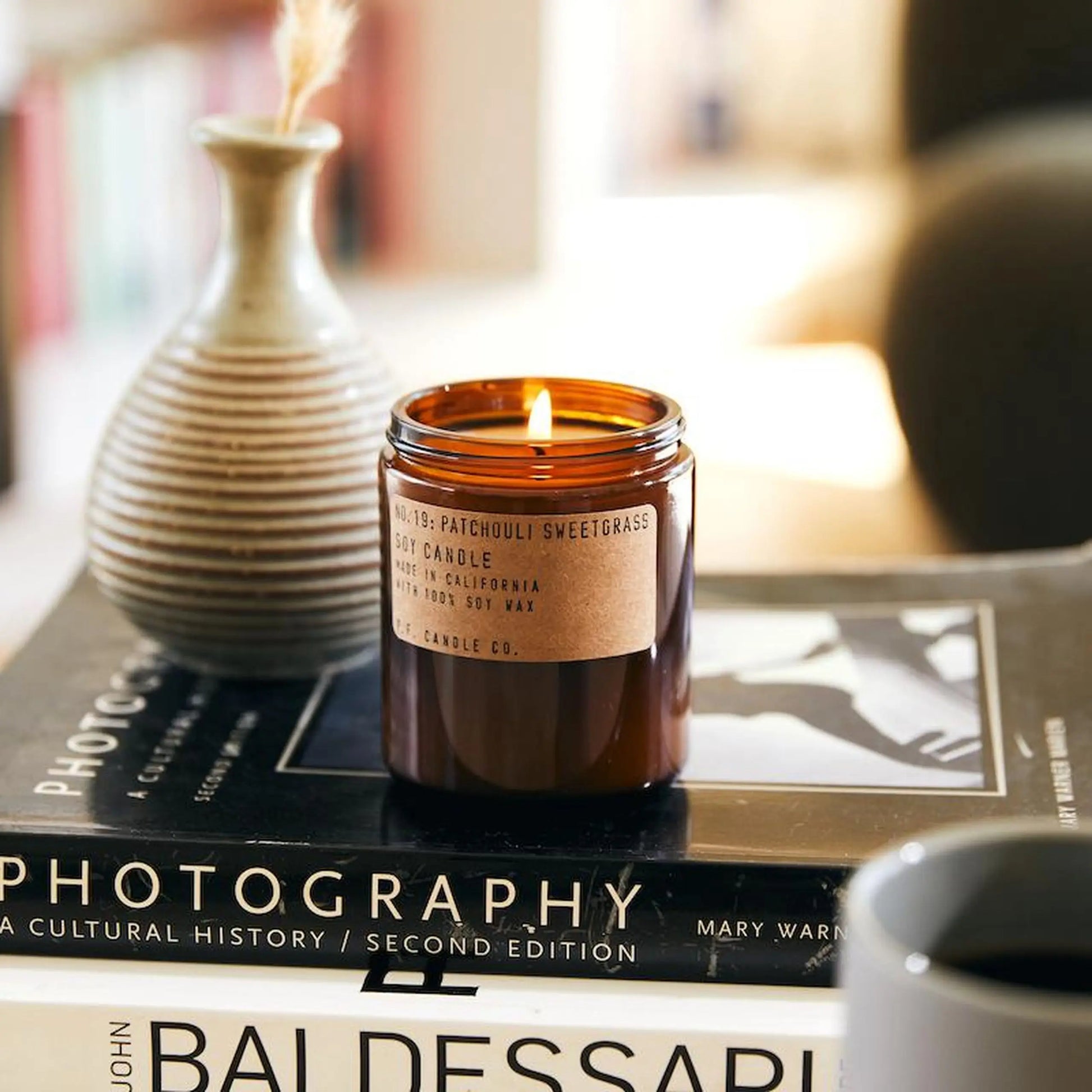 The P.F. Candle Co. No. 19 Patchouli Sweetgrass 7.2 oz Soy Candle, made with clean soy wax, rests on two art books next to a ceramic vase and coffee mug, creating a calm, inviting atmosphere in a softly blurred setting.