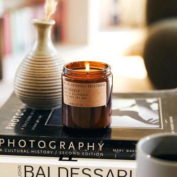 The P.F. Candle Co. No. 19 Patchouli Sweetgrass 7.2 oz Soy Candle, made with clean soy wax, rests on two art books next to a ceramic vase and coffee mug, creating a calm, inviting atmosphere in a softly blurred setting.