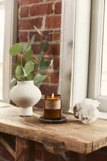 A white ceramic vase with eucalyptus branches, a lit P.F. Candle Co. No. 21 Golden Coast 7.2 oz Soy Candle, and a white crystal cluster rest on a rustic wood table by an exposed brick window, evoking fresh coastal vibes.
