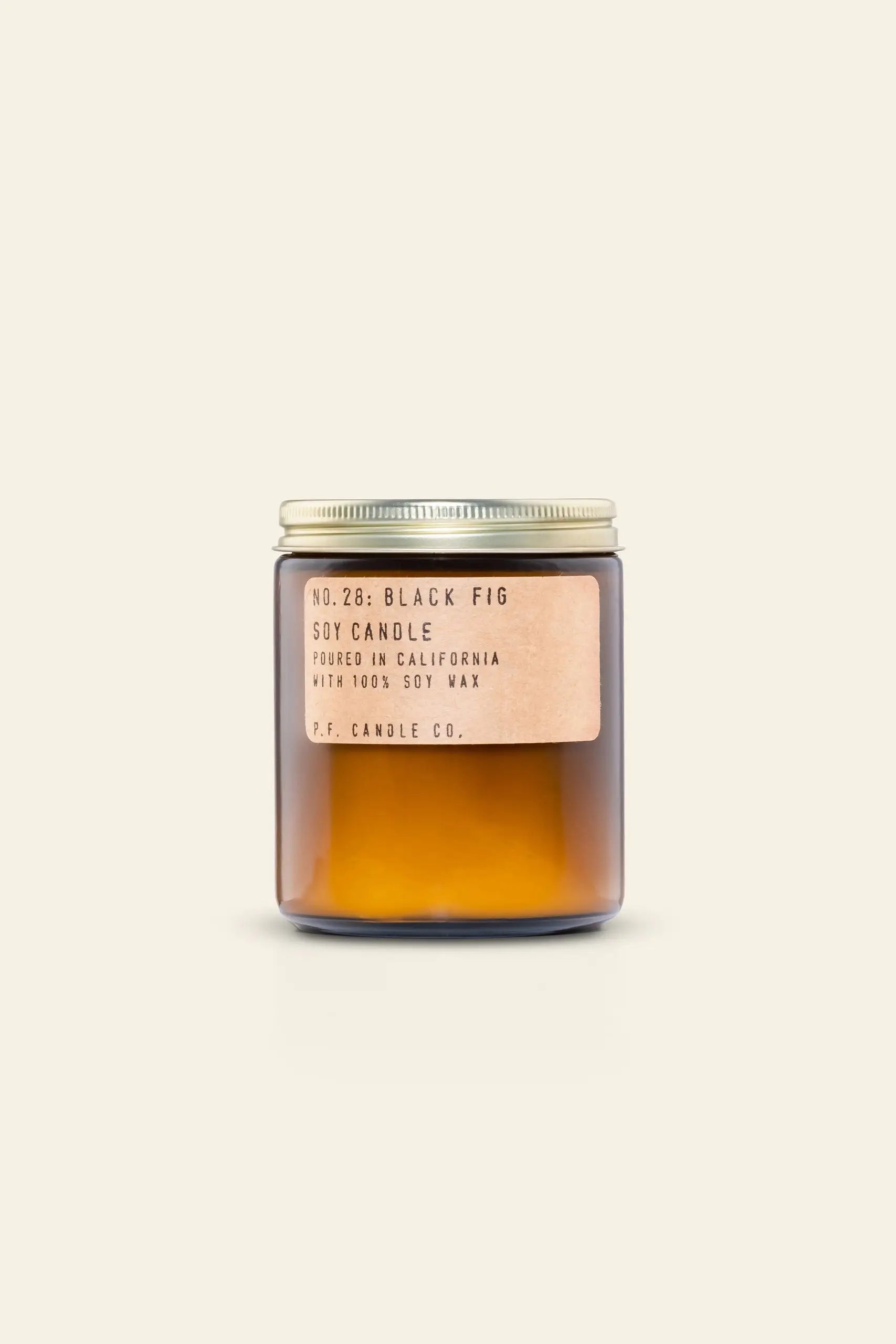 The P.F. Candle Co. No. 28 Black Fig 7.2 oz Soy Candle features a brown glass jar with a gold lid and tan label, hand-poured in California with 100% soy wax, and evokes the cozy scent of a winter fireplace against a beige background.