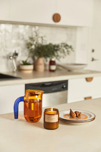 A lit P.F. Candle Co. No. 28 Black Fig 7.2 oz Soy Candle, a glass mug with orange liquid and blue handle, and a plate with two dates rest on a kitchen counter; blurred kitchen elements appear in the background.