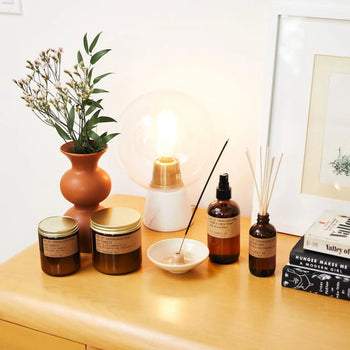 A wooden dresser displays a P.F. Candle Co. No. 32 Sandalwood Rose 7.2 oz Soy Candle, glass jars, a brown vase with flowers, a glowing lamp, incense in a dish, reed diffusers, stacked books, and a framed picture on a white wall.