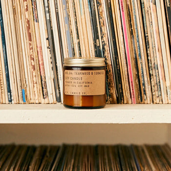 A P.F. Candle Co. No. 4 Teakwood & Tobacco 3.5 oz Mini Candle rests on a white shelf in front of vinyl records with colorful spines.