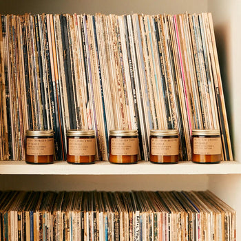 A shelf displays vinyl records, with five P.F. Candle Co. No. 4 Teakwood & Tobacco 3.5 oz Mini Candles featuring tan labels lined up in front on the middle shelf, and more records stored on the lower shelf.